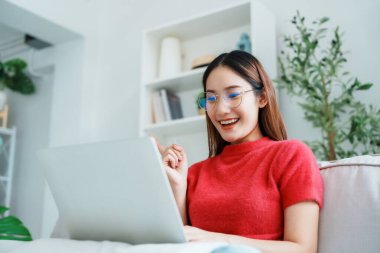 Evdeki kanepede otururken dizüstü bilgisayar kullanan mutlu genç Asyalı kadın.