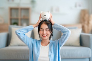 Genç Asyalı kadın domuz kumbarasını tutuyor ve evde yerde oturuyor. Para ve mali yatırım tasarrufu