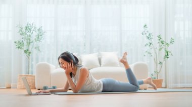 Genç Asyalı bir kadın laptopun önünde yoga minderinin üzerinde yerde yatıyor ve evde antrenman yapıyor. Egzersiz yapmadan önce internetten antrenörle iletişim kuran güzel, gülümseyen bir kadın.