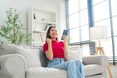 Oturma odasında komik mobil uygulamalar kullanarak rahat bir koltukta oturan gülümseyen genç Asyalı kadın. Evdeki Kadın, Çevrimiçi Alışveriş Yapıyor, Arkadaşlarına Mesajlaşıyor, Sosyal Medya 'ya Gönderiyor