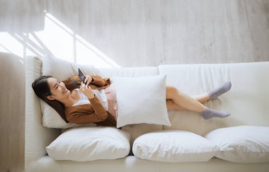 Mutlu, neşeli, mutlu, pozitif Asyalı bir kadının telefondaki görüntüsü. Uygulama kullanarak gülümseyen bir kız. İnternette akıllı telefondan kanepeye uzanmış.
