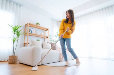 Ev temizliği ve eğlence. Genç, tapılası Asyalı kadın oturma odasını temizliyor, mikrofon olarak paspası kullanıyor, ev işlerini seviyor.