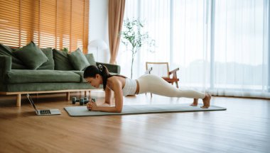 Spor kıyafetli genç bir Asyalı kadın oturma odasında dizüstü bilgisayarla antrenman dersleri izlerken antrenman minderinde tahta döşeme yapıyor. Spor ve eğlence konsepti