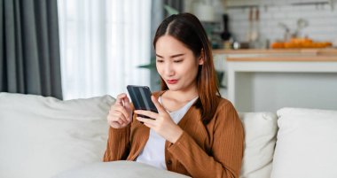 Oturma odasında komik mobil uygulamalar kullanarak rahat bir koltukta oturan gülümseyen genç Asyalı kadın. Evdeki Kadın, Çevrimiçi Alışveriş Yapıyor, Arkadaşlarına Mesajlaşıyor, Sosyal Medya 'ya Gönderiyor