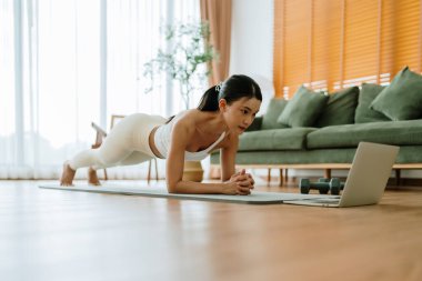 Spor kıyafetli genç bir Asyalı kadın oturma odasında dizüstü bilgisayarla antrenman dersleri izlerken antrenman minderinde tahta döşeme yapıyor. Spor ve eğlence konsepti