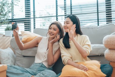 İki genç Asyalı kadın gülümseyip mutlu olurken evlerindeki oturma odalarında fotoğraf çektirip kameraya bakıyorlar.