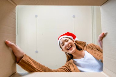 Şaşırmış Asyalı genç bir kadının Noel şapkası takıp bavullarını boşaltması. Kutuyu açıp içine bakmak. Paket kutusu, teslimat servisi. İnsan duyguları ve yüz ifadesi