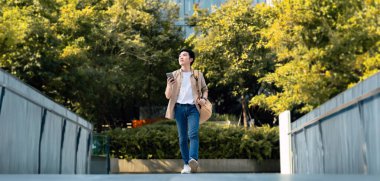 Akıllı telefon kullanan yakışıklı Asyalı öğrencinin portresi. Genç bir adam elinde cep telefonuyla mutlu bir şekilde dışarıda yürüyor.