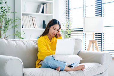 Genç Asyalı kadın evdeki koltukta otururken dizüstü bilgisayar kullanıyor.