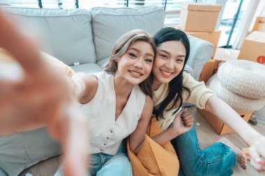 İki genç Asyalı kadın gülümseyip mutlu olurken evlerindeki oturma odalarında fotoğraf çektirip kameraya bakıyorlar.