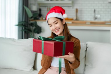 Genç Asyalı kadın Noel şapkası takıyor ve hediye kutusunu açıyor oturma odasında kanepede oturuyor. Mutlu yıllar, Şükran Günü, Mutlu Noeller