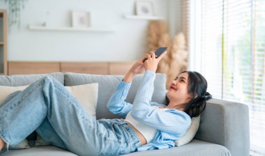 Mutlu, pozitif, genç, Asyalı bir kadın evde telefonda, gülümseyen bir kız. Uygulama kullanıyor ve internette akıllı telefonuyla kanepeye uzanıyor.