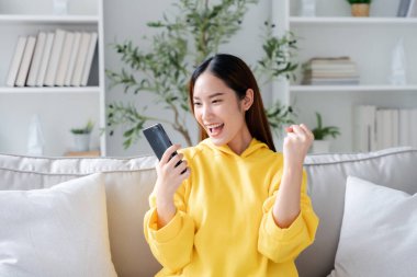 Oturma odasında komik mobil uygulamalar kullanarak rahat bir koltukta oturan gülümseyen genç Asyalı kadın. Evdeki Kadın, Çevrimiçi Alışveriş Yapıyor, Arkadaşlarına Mesajlaşıyor, Sosyal Medya 'ya Gönderiyor