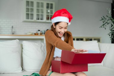 Genç Asyalı kadın Noel şapkası takıyor ve hediye kutusunu açıyor oturma odasında kanepede oturuyor. Mutlu yıllar, Şükran Günü, Mutlu Noeller