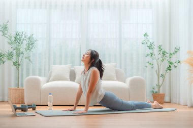 Genç Asyalı kadın oturma odasında bilgisayarlı dizüstü bilgisayarla antrenman yaparken yoga antrenmanını spor minderinde yapıyor.