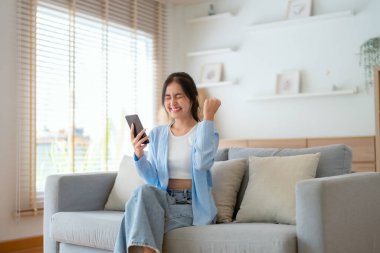 Oturma odasında komik mobil uygulamalar kullanarak rahat bir koltukta oturan gülümseyen genç Asyalı kadın. Evdeki Kadın, Çevrimiçi Alışveriş Yapıyor, Arkadaşlarına Mesajlaşıyor, Sosyal Medya 'ya Gönderiyor