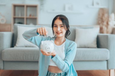 Genç Asyalı kadın domuz kumbarasını tutuyor ve evde yerde oturuyor. Para ve mali yatırım tasarrufu