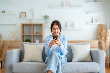 Oturma odasında komik mobil uygulamalar kullanarak rahat bir koltukta oturan gülümseyen genç Asyalı kadın. Evdeki Kadın, Çevrimiçi Alışveriş Yapıyor, Arkadaşlarına Mesajlaşıyor, Sosyal Medya 'ya Gönderiyor