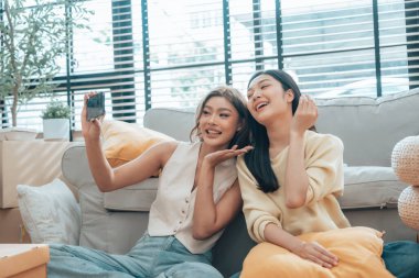 İki genç Asyalı kadın gülümseyip mutlu olurken evlerindeki oturma odalarında fotoğraf çektirip kameraya bakıyorlar.