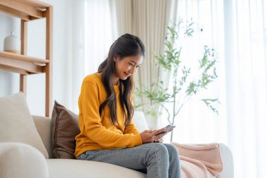 Oturma odasında komik mobil uygulamalar kullanarak rahat bir koltukta oturan gülümseyen genç Asyalı kadın. Evdeki Kadın, Çevrimiçi Alışveriş Yapıyor, Arkadaşlarına Mesajlaşıyor, Sosyal Medya 'ya Gönderiyor