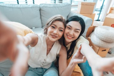 İki genç Asyalı kadın gülümseyip mutlu olurken evlerindeki oturma odalarında fotoğraf çektirip kameraya bakıyorlar.