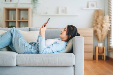 Mutlu, pozitif, genç, Asyalı bir kadın evde telefonda, gülümseyen bir kız. Uygulama kullanıyor ve internette akıllı telefonuyla kanepeye uzanıyor.