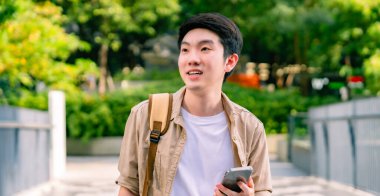 Akıllı telefon kullanan yakışıklı Asyalı öğrencinin portresi. Genç adam elinde cep telefonuyla mutlu bir şekilde dışarıda yürüyor.
