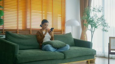 Oturma odasında komik mobil uygulamalar kullanarak rahat bir koltukta oturan gülümseyen genç Asyalı kadın. Evdeki Kadın, Çevrimiçi Alışveriş Yapıyor, Arkadaşlarına Mesajlaşıyor, Sosyal Medya 'ya Gönderiyor