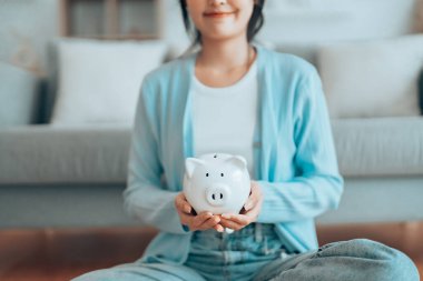 Genç Asyalı kadın domuz kumbarasını tutuyor ve evde yerde oturuyor. Para ve mali yatırım tasarrufu