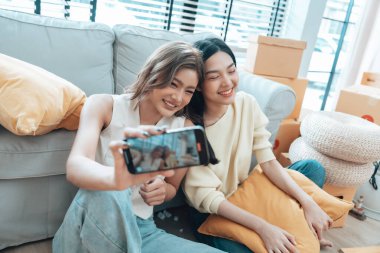 İki genç Asyalı kadın gülümseyip mutlu olurken evlerindeki oturma odalarında fotoğraf çektirip kameraya bakıyorlar.