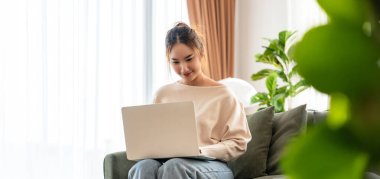 Oturma odasında dizüstü bilgisayarla çalışan mutlu genç Asyalı kadın. Serbest çalışan kadın kanepeye oturmuş Webinar 'ı izliyor, internetten ders çalışıyor.