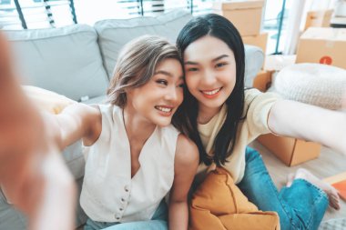 İki genç Asyalı kadın gülümseyip mutlu olurken evlerindeki oturma odalarında fotoğraf çektirip kameraya bakıyorlar.