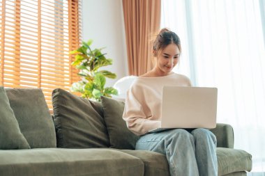 Oturma odasında dizüstü bilgisayarla çalışan mutlu genç Asyalı kadın. Serbest çalışan kadın kanepeye oturmuş Webinar 'ı izliyor, internetten ders çalışıyor.