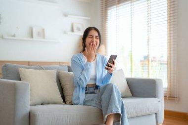 Oturma odasında komik mobil uygulamalar kullanarak rahat bir koltukta oturan gülümseyen genç Asyalı kadın. Evdeki Kadın, Çevrimiçi Alışveriş Yapıyor, Arkadaşlarına Mesajlaşıyor, Sosyal Medya 'ya Gönderiyor