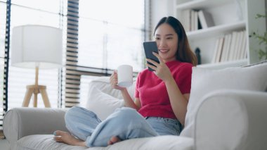 Evde kanepede otururken cep telefonu kullanan çekici, gülümseyen Asyalı kadın.
