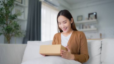Mutlu genç Asyalı kadın teslimat paketini evde açıyor. Güzel bayan müşteri kutusunu açar ve oturma odasında kanepede oturur.