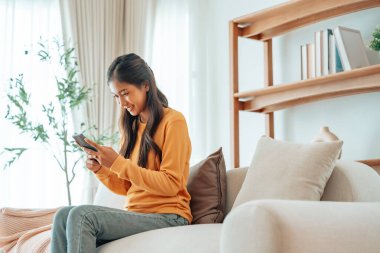 Happy young asian woman relax on comfortable couch at home texting messaging on smartphone, smiling girl use cellphone, chatting online message, shopping online from home