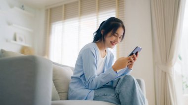 Oturma odasında komik mobil uygulamalar kullanarak rahat bir koltukta oturan gülümseyen genç Asyalı kadın. Evdeki Kadın, Çevrimiçi Alışveriş Yapıyor, Arkadaşlarına Mesajlaşıyor, Sosyal Medya 'ya Gönderiyor