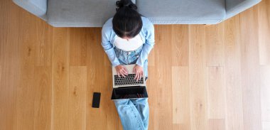 Asya 'lı genç bir kadının oturma odasında cep telefonuyla bilgisayarda çalışırken en iyi görüntüsü.