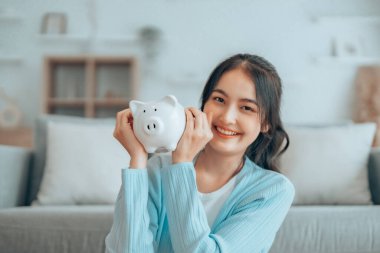 Genç Asyalı kadın domuz kumbarasını tutuyor ve evde yerde oturuyor. Para ve mali yatırım tasarrufu