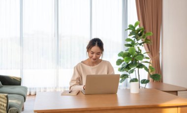 Oturma odasında dizüstü bilgisayarla çalışan mutlu genç Asyalı kadın. Serbest çalışan kadın masa başında oturmuş Webinar izlerken notlar yazıyor, internetten ders çalışıyor.