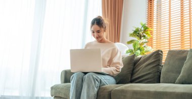 Oturma odasında dizüstü bilgisayarla çalışan mutlu genç Asyalı kadın. Serbest çalışan kadın kanepeye oturmuş Webinar 'ı izliyor, internetten ders çalışıyor.