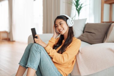 Evdeki oturma odasında kulaklık ve cep telefonuyla müzik dinleyen Asyalı bir kadın. Motivasyon ve gülümseme için şarkı, radyo, podcast ve kulaklık.
