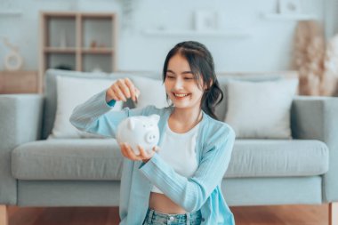 Genç Asyalı kadın domuz kumbarasını tutuyor ve evde yerde oturuyor. Para ve mali yatırım tasarrufu