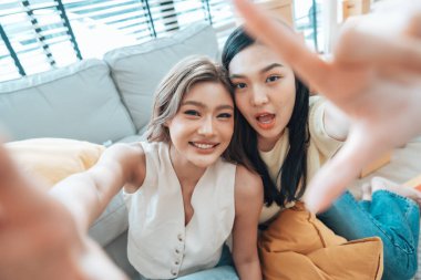 İki genç Asyalı kadın gülümseyip mutlu olurken evlerindeki oturma odalarında fotoğraf çektirip kameraya bakıyorlar.