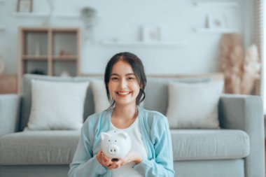 Genç Asyalı kadın domuz kumbarasını tutuyor ve evde yerde oturuyor. Para ve mali yatırım tasarrufu