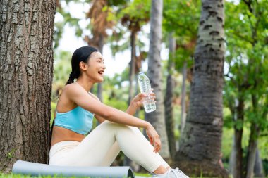Aktif giysili genç bir Asyalı kadın bir ağacın altında oturmuş parkta spor yaptıktan sonra su içiyor. Rahatlamış ve rahatlamış görünüyor. Açık havada huzurlu bir ortamda susuzluktan ve doğadan hoşlanıyor.