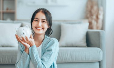 Genç Asyalı kadın domuz kumbarasını tutuyor ve evde yerde oturuyor. Para ve mali yatırım tasarrufu