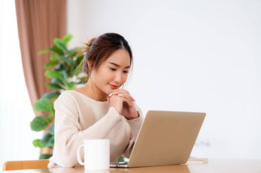 Oturma odasında dizüstü bilgisayarla çalışan mutlu genç Asyalı kadın. Serbest çalışan kadın masa başında oturmuş Webinar izlerken notlar yazıyor, internetten ders çalışıyor.
