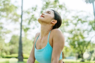 Genç Asyalı kadın güneşli bir günde arka planda ağaçlarla birlikte temiz hava soluyor. Sağlık, sağlıklı yaşam tarzı, farkındalık ve açık hava sporu kavramı.